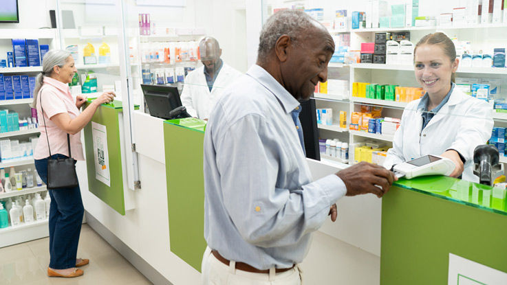 At a pharmacy counter, two friendly young pharmacists are each helping an older customer with their prescriptions.