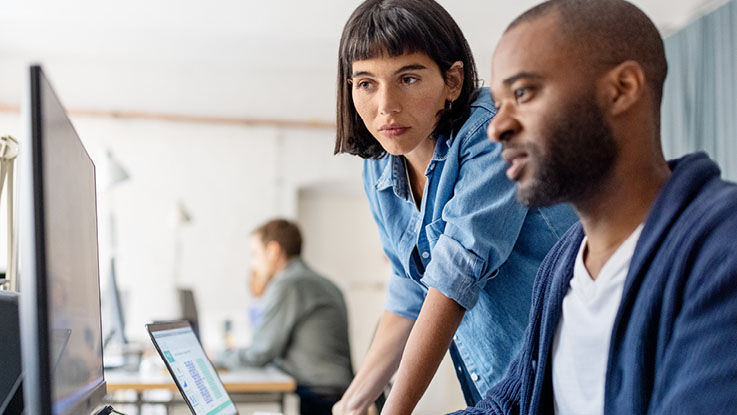 In an office, two coworkers look at information together on a computer as they work to solve a problem.
