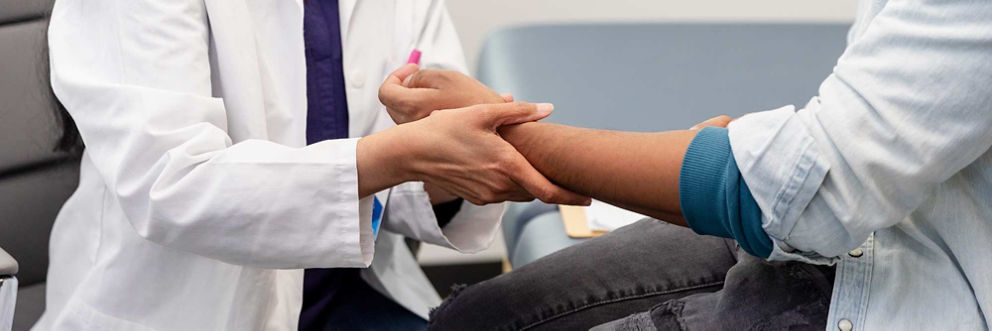 A doctor and patient sit side-by-side while the doctor examines the patient's forearm.