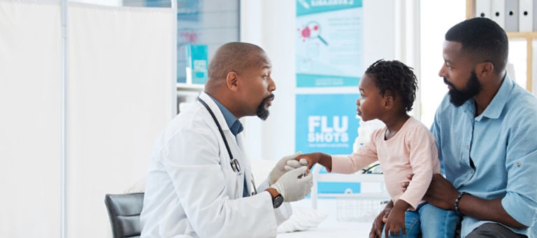 In a doctor’s office, a doctor smiles as he speaks to a young child who is sitting on his father’s lap