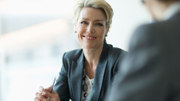 A woman wearing business attire smiles as she speaks with a colleague