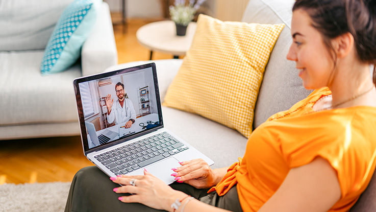 A woman sits on a couch with a laptop on her lap during a video visit with a doctor.