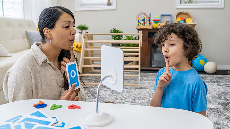 In an friendly office with children's toys on a table, a speech therapist shows a child how to pronounce vowel sounds.