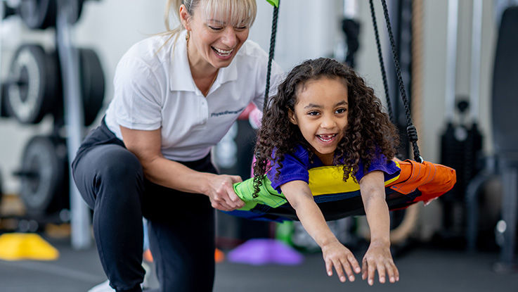 A girl lays down on a swing while an occupational therapist pushes her.