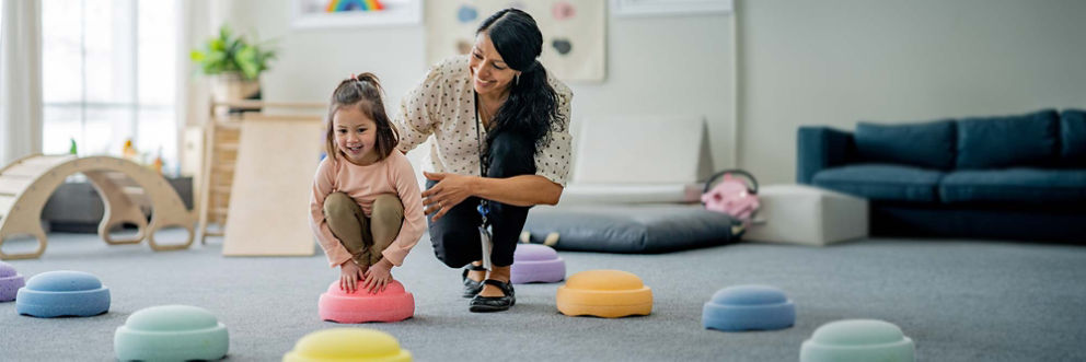 A child squats on a colorful cushion while her therapist guides her through a game that improves balance and coordination.