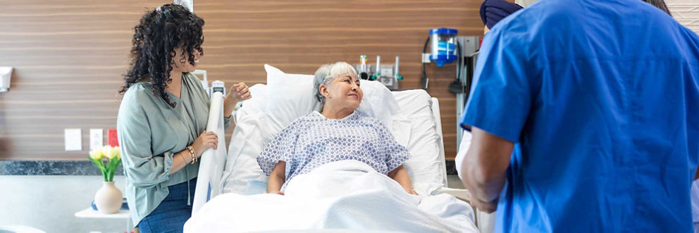 An older woman in a hospital bed smiles towards her care team while her daughter stands at her side.