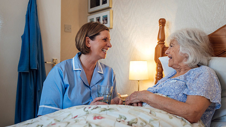 A hospice nurse offers a glass of water to a smiling older woman who’s in bed.