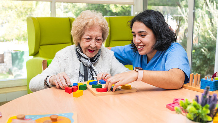 A woman in scrubs helps an elderly woman with a block puzzle.