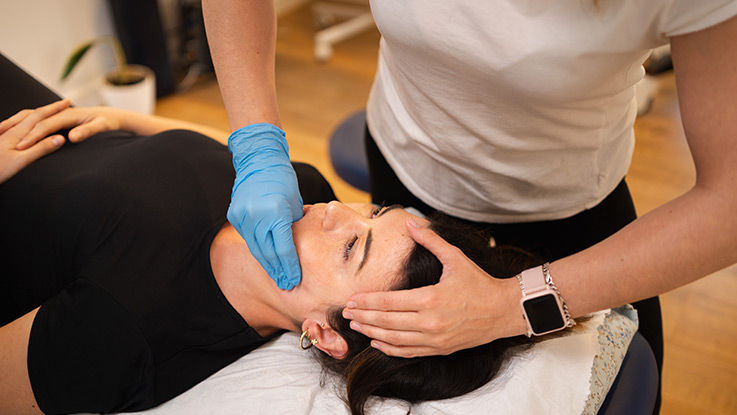 A patient reclines on an examination table while a physical therapist massages her jaw.