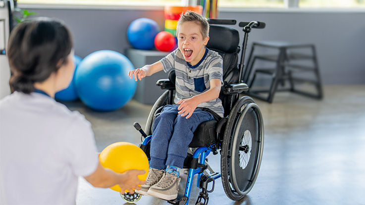 A boy in a wheelchair plays catch with a physical therapist.