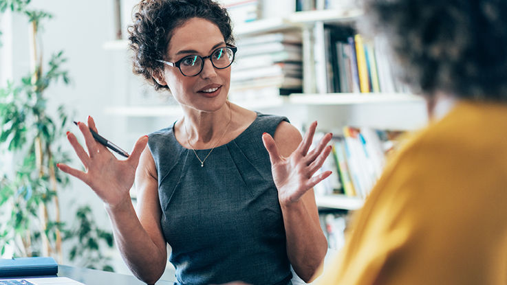 Two smiling businesswomen chat together, one with her hands open and raised to demonstrate a points