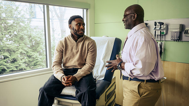 A patient sits on an examination table and talks with his doctor.