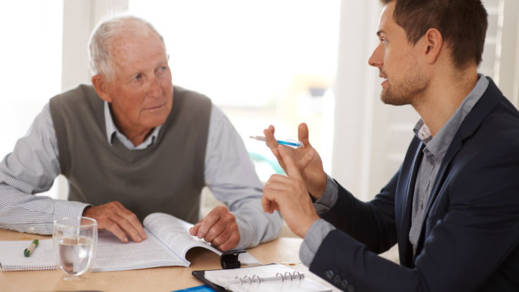 Sitting at his dinner table, an older man listens as his Medicare broker discusses plan details.