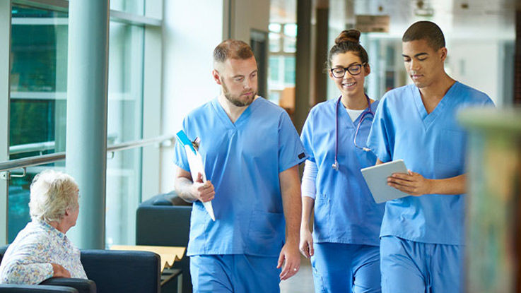 Three doctors walk down a health care clinic hallway and talk with each other while referring to information on a tablet computer one of them is holding