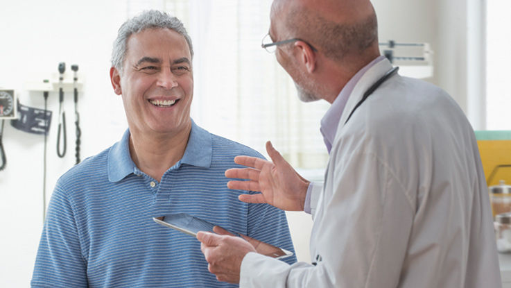 In an exam room, a man smiles as his doctor talk about his health