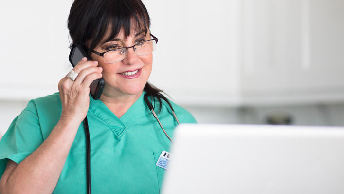 A nurse smiles as she talks on the phone and looks up information on a computer