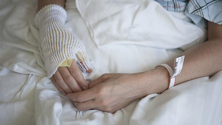 A close-up of a patient's hands in a hospital bed. One hand is covered in a bandage while they heal.