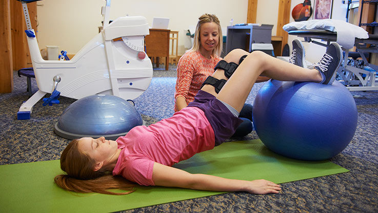 A physical therapist guides a patient through a pelvic bridge exercise with the help of an yoga ball.
