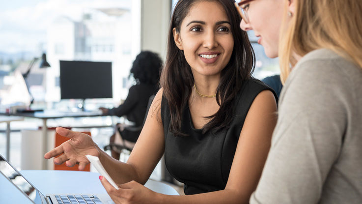 Two women sitting at a desk in front of an open laptop.