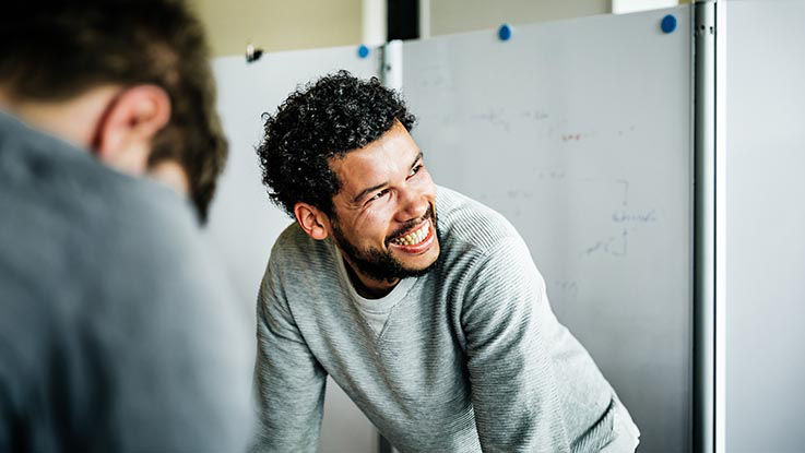 A man standing in front of a whiteboard smiles as he looks at a colleague