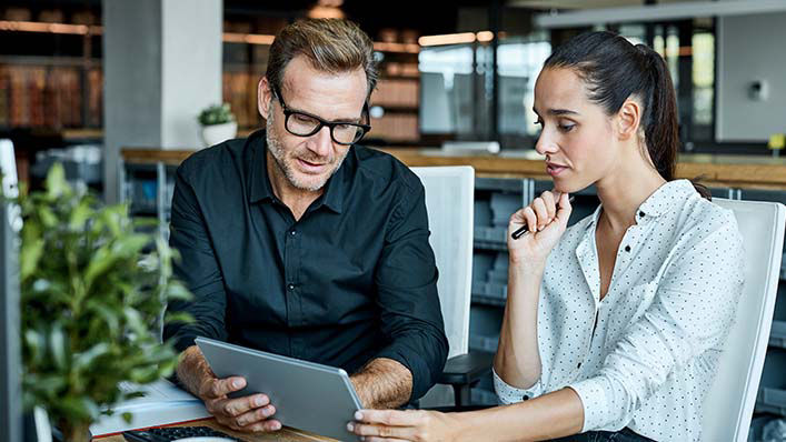 Sitting at an office desk, a man describes information on a tablet to his female coworker.