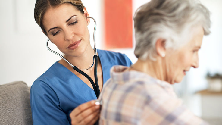 An older woman sits in her living room, having her lungs examined by a young female nurse with a stethoscope.