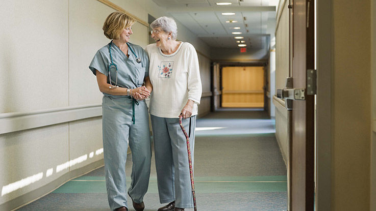 A transitional care specialist helps an elderly woman use a cane to walk the hospital hallways.