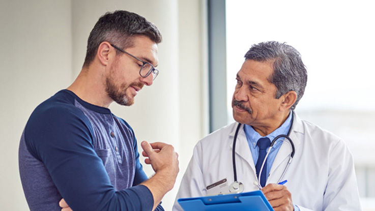 In a doctor’s office, a man speaks with a doctor while the doctor listens and takes notes on a clipboard
