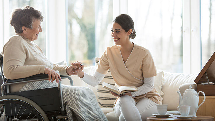 A nurse with a book in her lap holds hands with an older woman in a wheelchair.