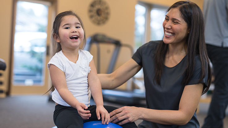 A child smiles as her therapist guides her through an exercise on a balance ball.