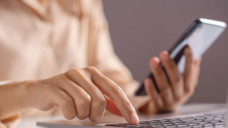 A woman refers to her phone as she enters information into her laptop