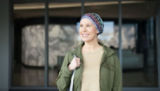 Smiling woman with head covered in a scarf stands in front of a building.