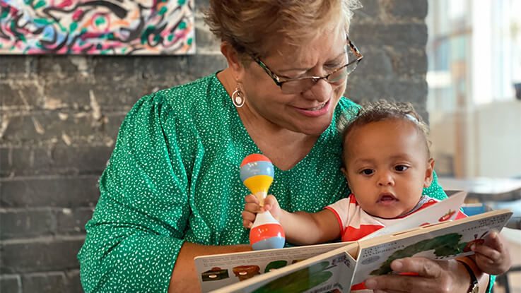 An older woman reads a picture book to her infant grandchild in her lap.