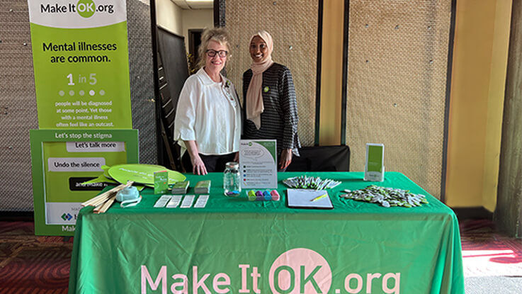 Two mental health professionals pose for a photo standing behind the Make It Ok informational booth.