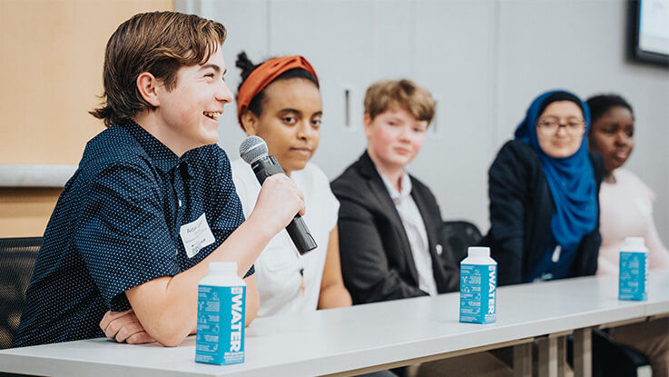 A teenage boy speaks into a microphone while sitting with other teens on the Teen Leadership Council.