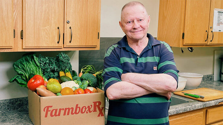 Tom Rabatin poses for a photo next to a box labeled FarmboxRx that is overflowing with fresh produce.