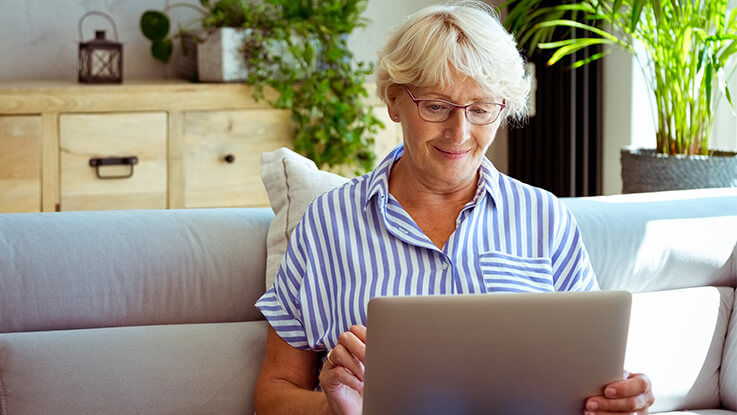 An older woman sits on the couch at home and researches expected costs of care on her laptop.