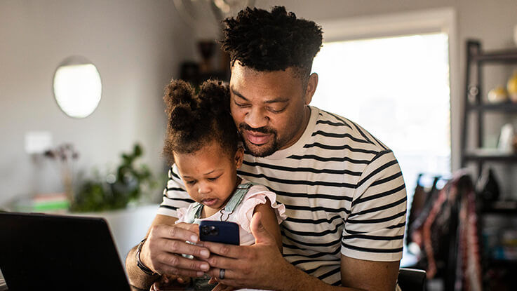 A father holding his toddler daughter in his lap uses his phone to access Save My Spot for same-day care.