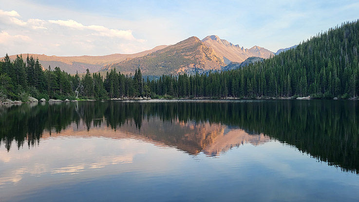 A photo titled “Nature’s perfect mirror” by Dom Ehman, capturing a forest and mountain reflected in a still lake.
