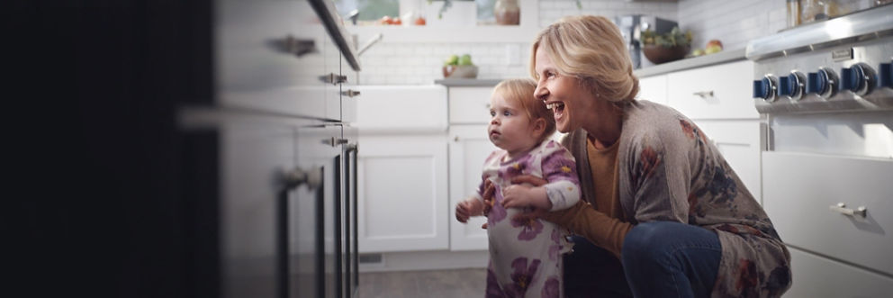 A laughing woman is crouched in a kitchen with her hands supporting a toddler. The toddler is looking intently at a drawer.